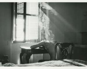 Black and white photograph of nude male figure slumped over a desk in a bedroom before a window with light and smoke streaming in