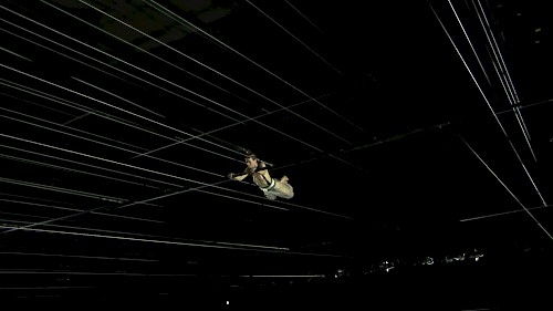 A photograph of a dark room and a person swinging from trapeze in the center of the picture