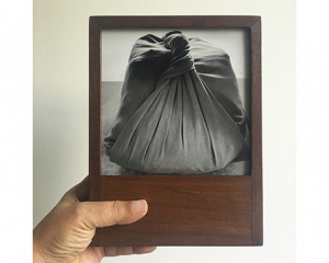 A hand holds a teak wood frame with a black and white photograph of a pothi, a knotted package made of fabric.