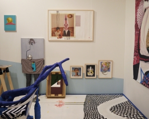 An installation photograph of a portion of Callicoon Fine Art's booth. There are 4 framed collages, a complex floor painted with small black triangles, and a blue sculpture attached to a chair. There are also two paintings near the left edge of the image that are figurative.