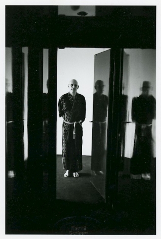 A black and white portrait of Michel Foucault in a robe, standing in a doorway. There is light in the hallway, his hands are behind his back.