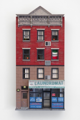 A sculpture made of foam and paper that depicts a building facade with a laundromat on the first floor, and apartments on the 3 floors above it. Half the building is red brick; the other half is beige stone.