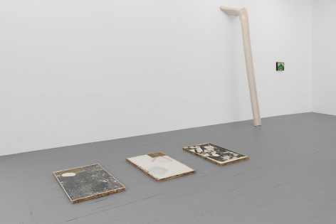 A photograph of the left half of the gallery. There are three collograph prints installed on the ground in short OSB platforms. There is a sculpture leaning against the wall, and a small abstract painting to the right of it. The painting is mostly green.