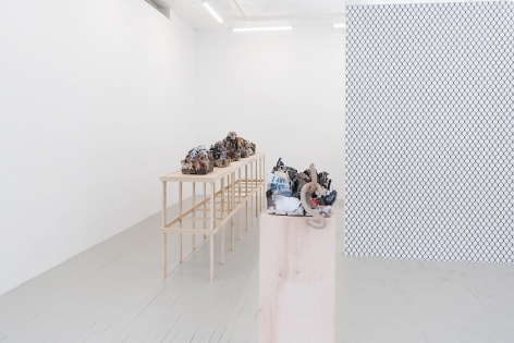 A photograph of the chainlink fence wallpaper at right, jutting to the left from the right. There is a raw wood pedestal with a single ceramic sculpture, and a long raw wood platform with a single row of 5 ceramic sculptures.