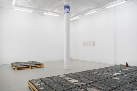 Installation image that shows 4 white tiles in the distance, an artwork on the floor made of black clay, and half of the large ceramic sculpture installed on the floor in the foreground
