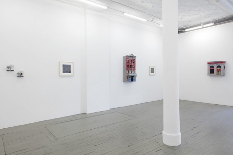 A photograph of the gallery interior, with 2 small sculptures at left installed on the wall. These works are followed by a black and white drawing, then a building facade sculpture, then another drawing that is illegible, then another facade sculpture on the opposite wall in the background.