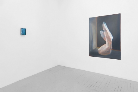 A view of 2 artworks around the corner of the gallery. On the left is a small abstract painting in blue tones; on the right is a kneeling naked woman seemingly made of cellophane, holding a mask of a human face. We see her shadow on the wall she's facing, with a black wall behind her, outlined in a warm yellow hue.