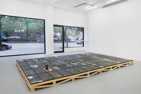 An installation image of Kahlil Robert Irving's large black clay tile artwork, installed on the floor on a wooden pedestal, with the windows of the gallery in the distance