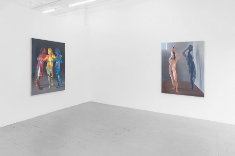 A photograph depicting two large paintings: one with three figures standing together, painted in red, yellow, and blue; the other with a single female naked body flattened like a paper doll, folding over herself, with an identical grey silhouette to the right of her.