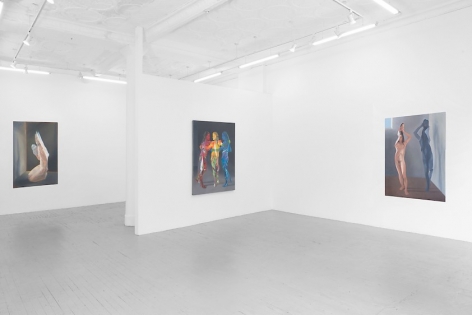 A photograph of the three large paintings: one of a single female figure not unlike a paper doll, folding over herself, at right; a painting of three figures in red, yellow, and blue on the temporary wall; and a painting of a naked female figure kneeling, looking like she's made of cellophane.