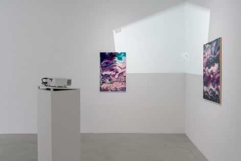 Two artworks are seen facing each other around the corner of the gallery. In the foreground is a projector on a white pedestal. On the wall on the corner is a white light, which is a video from Google Maps projecting the interior of the gallery onto itself.