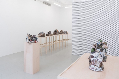A photograph that partially captures the square raw wood table, and we see 2 ceramic sculptures, blurring together somewhat. There is a single raw wood pedestal in the middle ground, and a long table at left, along the wall. On the long table are 5 sculptures. At right in the middle ground we see the chainlink fence wallpaper on the gallery's temporary wall.
