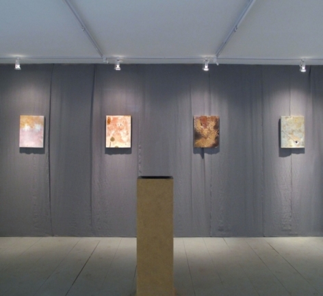 A photograph of 4 works on a gray wall, and a wooden pedestal in the middle