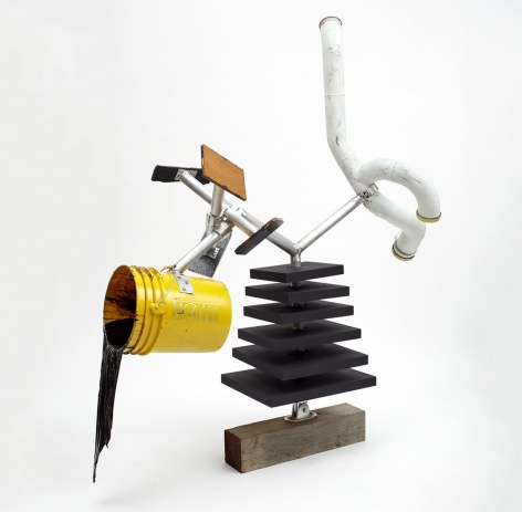 A mixed-media sculpture: wood block base, with 6 layers of wood slabs painted black above it. From the slabs there are 2 aluminum arms: one ends in a yellow bucket with black resin flowing out, froze, and the other is a white tube with 3 openings