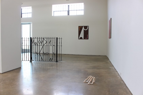 A photograph of the installation at SITE 131 that includes a fence by A.K. Burns in the background at left, 2 paintings by Ulrike Müller installed around the central and right wall (on either side of the corner), and Lee Relvas wooden shoes in the foreground.