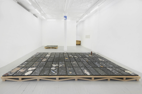 An installation image of the entire gallery, including 2 black clay tiled pieces installed on the floor (large in the foreground; smaller in the background) as well as a swatch of blue sky adhered to the top of a white concrete column in the center of the room