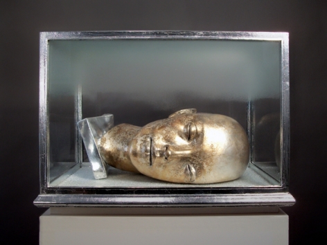 A photograph of a golden head in a vitrine, laying on its side with no hair, eyes closed