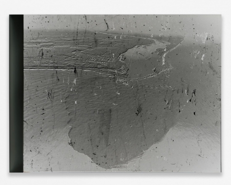 A grey photograph of an aerial view of the BP Oil Spill in the ocean, with small flecks upon the surface. The photograph is mounted on black plexiglass.