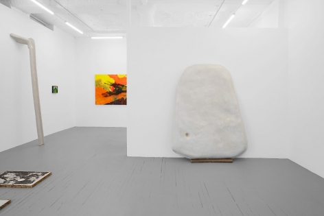 A photograph of the gallery where we see the temporary wall with a large white sculpture upon it. There is a partial view of a colorful painting on the back wall and a smaller painting at left of that. There is a large sculpture leaning against the wall at left, and a print installed on the ground near the left-corner of the photograph.