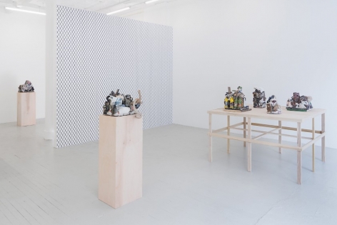 A photograph of the main area of the gallery: at right is a square raw platform with 4 ceramic sculptures upon it. On the temporary gallery wall is a wallpaper visualizing a chainlink pattern in black and white. There are also 2 raw wood pedestals with one sculpture on them, respectively.