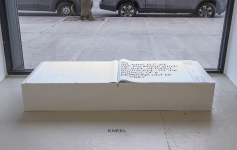 A book, black text on white pages, installed on a custom white pedestal in front of a window, with instructions on the floor to KNEEL